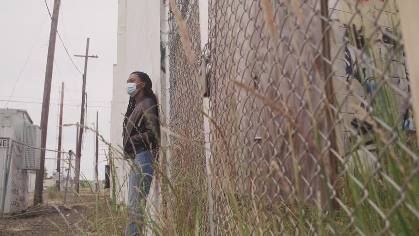 A woman wearing a medical mask stands in an alley with her back against a graffitied building during the day