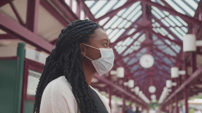 Woman with long hair braids wearing face mask stands in large building with high glass ceiling