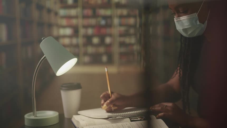Woman with braids and a mask writes in a notebook in the library with bookshelves in the background