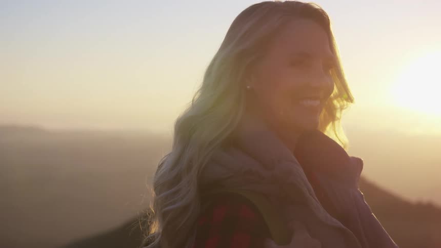 Blonde woman with a plaid shirt and a brown vest smiles brightly on the mountain at sunset