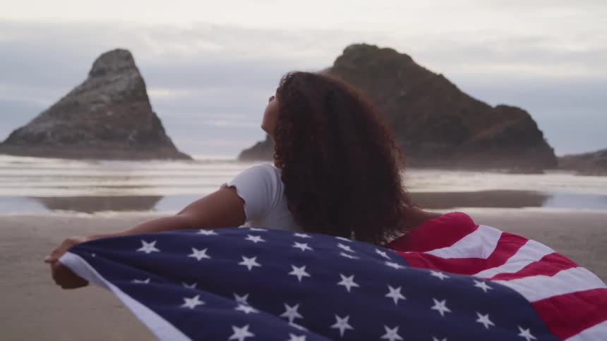 A girl is running across a beach carrying an american flag behind her