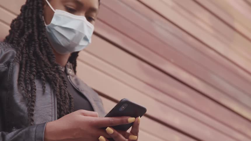 Young woman wearing a surgical face mask using a smartphone near a wall