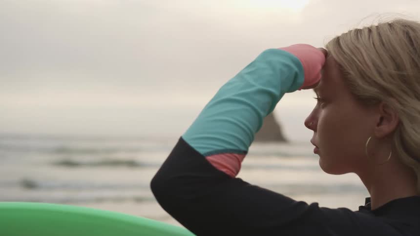 Woman holding surfboard looks out to sea as dog stands in background