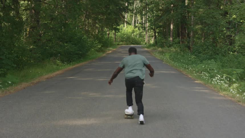 A man rides a skateboard down a road through a forest during the day