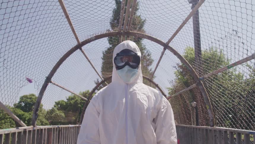A person with goggles and a mask and a hazmat suit standing in an overpass with trees the sky and clouds in the background