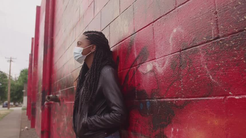 A woman with a mask standing by a wall with trees the sky and clouds in the background