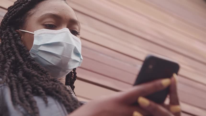 A woman in a medical mask uses her smartphone while leaning against a wall outdoors during the day