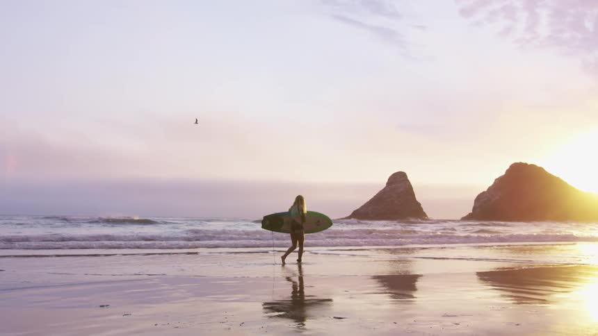 A young woman walks slowly along the beach while holding a surfboard under her arm
