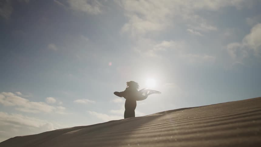 The silhouette of a woman wearing a gown with a sheer veil billowing around her as she walks on the ridge of a sand dune