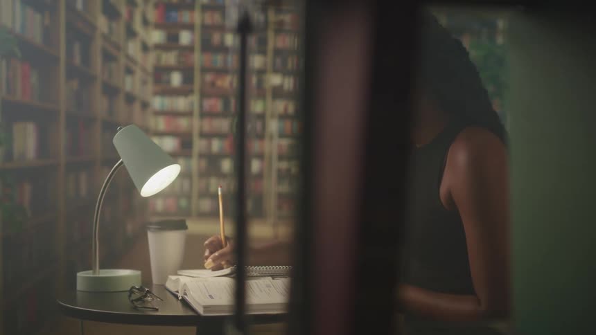 Woman with braids and a mask writes in a notebook in the library with bookshelves in the background
