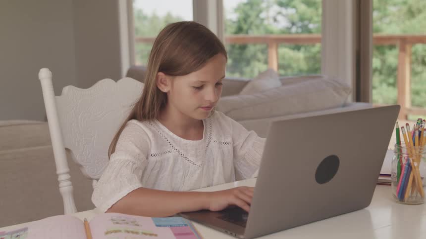 A girl sitting at a desk at home typing ona laptop with her workbook and pencil next to her
