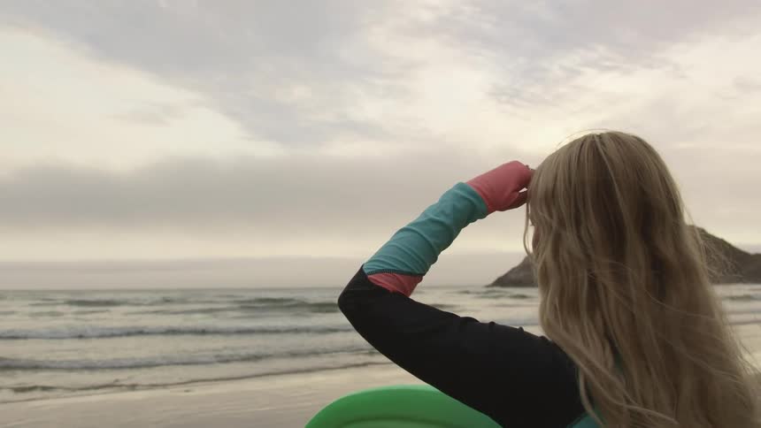A female surfer shields her eyes from the sunlight as she looks out over the ocean from a beach while holding a surfboard on a cloudy evening