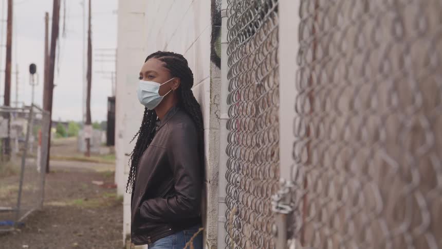 Woman wearing face mask leans against building wall near street intersection