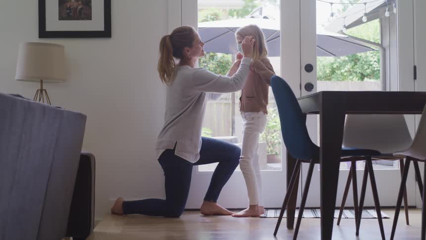 A mother kneeling to put a face mask on her daughter at home