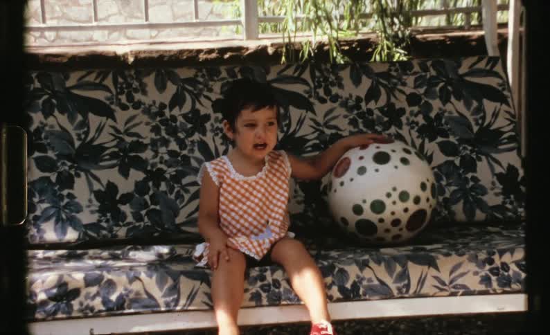 A little girl sitting on a chair with a ball with a bicycle in the foreground and a wall in the background