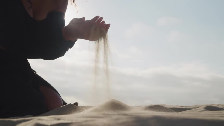 A woman scoops sand from the ground before letting it slip through her fingers as she kneels in the sands of a desert during the day