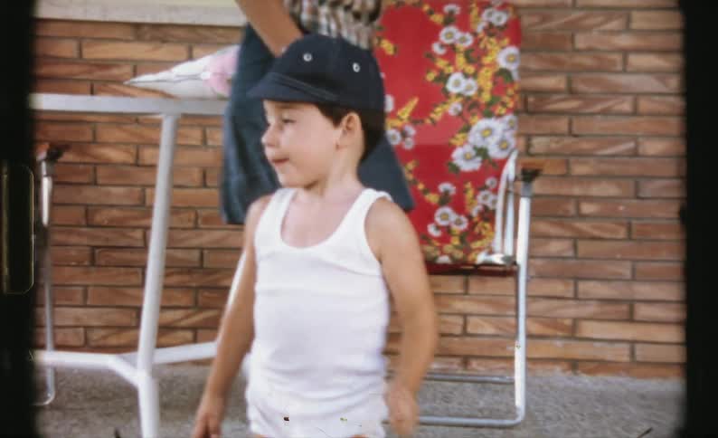 A boy and girl look into the camera while they pace back and forth on the porch of a house during the day