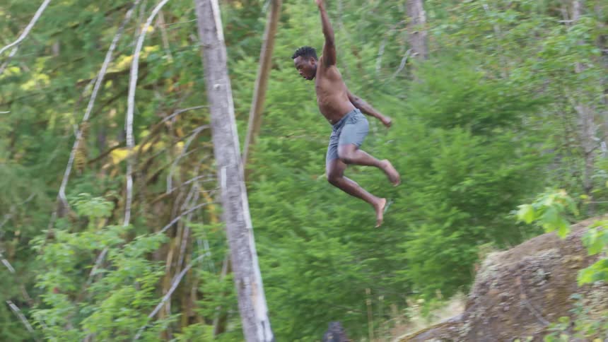 Strong muscular shirtless man jumping off the edge of a cliff into a lake in the forest