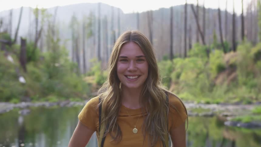 A woman stands smiling in front of a pond with trees and hills behind her