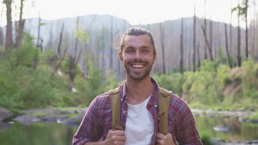 A man with a backpack smiling with a creek trees the sky and clouds in the background