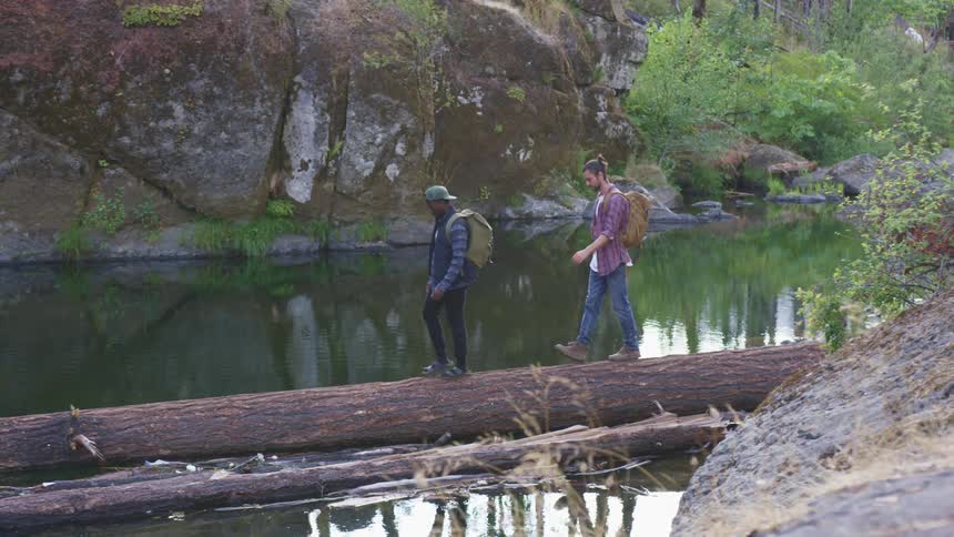 Two men with backpacks walking on a log over a creek