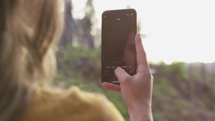 A woman with a smart phone taking a picture of a lake with the sky and clouds in the background