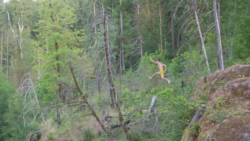 A man running and jumping off a cliff into a creek with trees the sky and clouds in the background