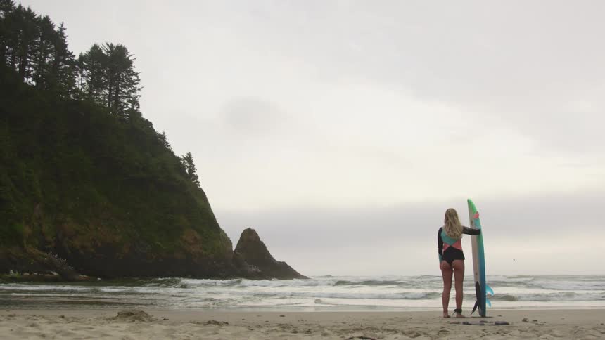 A woman holding a surfboard standing on a beach with the ocean sky and clouds in the background