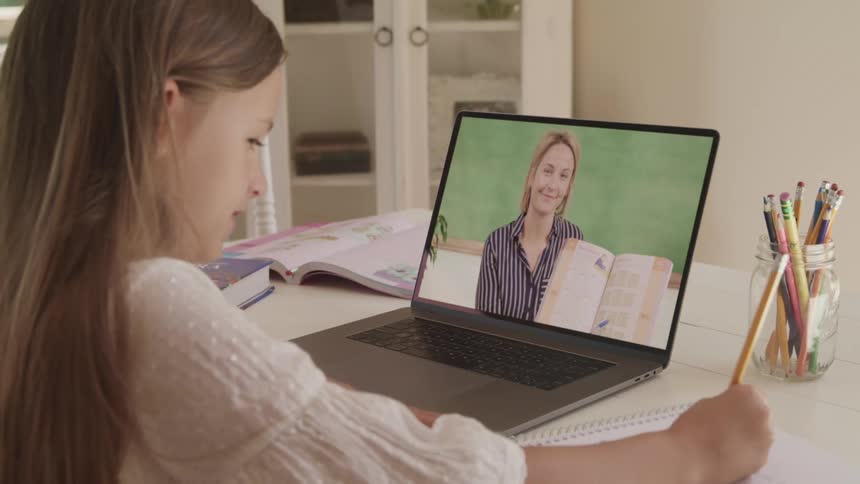 A girl writes in a notebook while being instructed by a woman through a video conference on a laptop