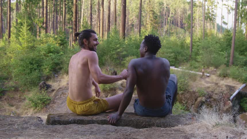 Two men sitting and shaking hands with dirt plants and trees in the background