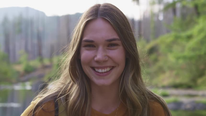 Young woman standing in front of a lake in the forest looks at the camera smiling