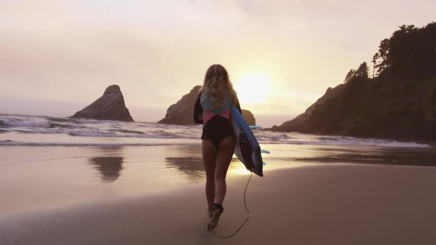 A woman running on a sandy beach with a surfboard at sunset