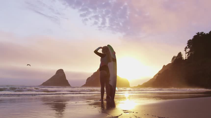 Young blonde woman wearing a colorful wet suit holding a green surfboard on the coast of a beach at sunset
