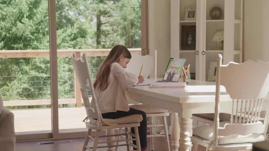 A girl sitting on a chair at a table looking at a laptop and writing on a paper with a pencil