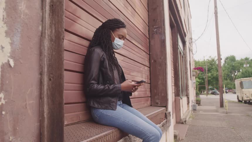 Woman with braids a mask a black leather jacket and jeans uses a smartphone in the street