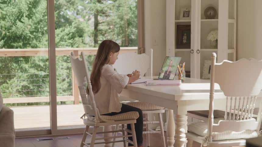 A young girls sits at a table in front of her laptop and waves to her distance learning instructor