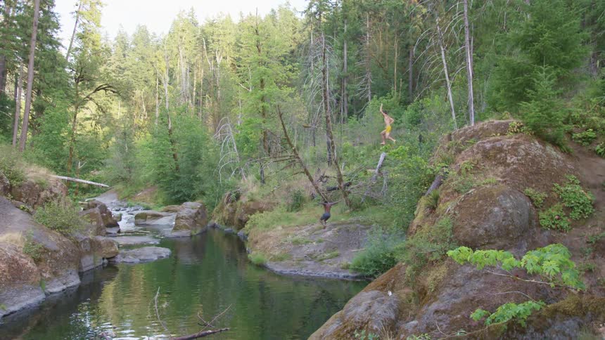 Two young shirtless men jumping into a river in the forest