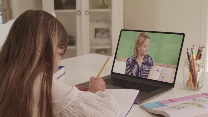 A young girl sits at a table with a laptop doing virtual school with a teacher on the screen
