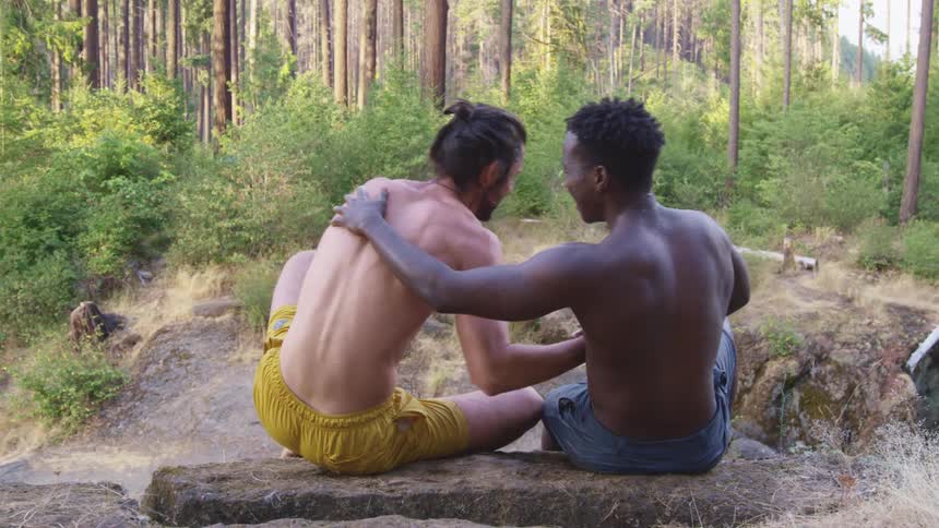 Two men shaking hands and hugging with plants trees and dirt in the background