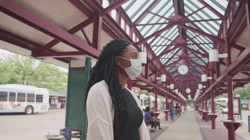 A woman with a mask standing in a bus station with a bus and trees in the background