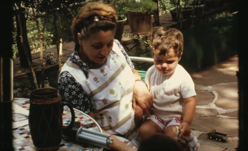 A woman sitting on a chair with a boy on her knee next to a table with plants in the background