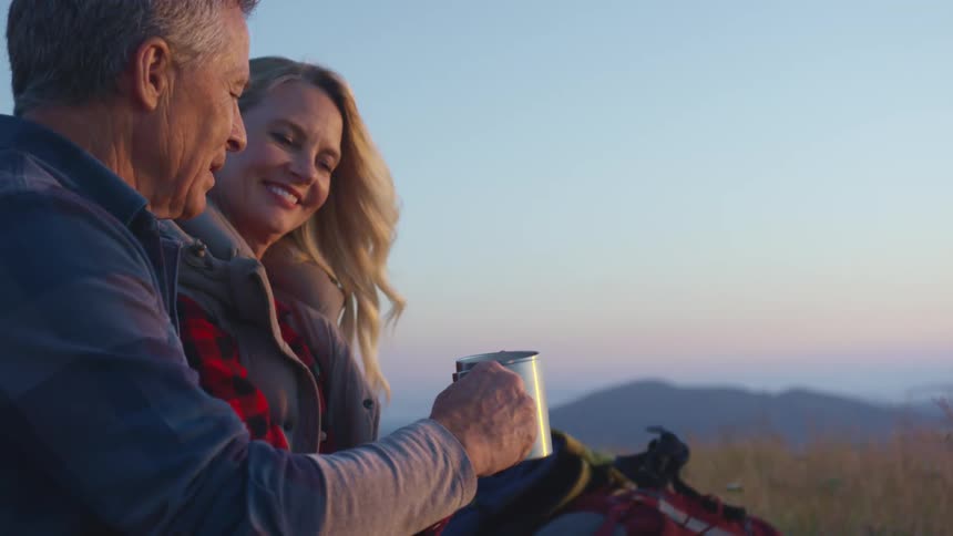 A man and woman toast with coffee mugs while sitting in the grass overlooking mountains at sunset