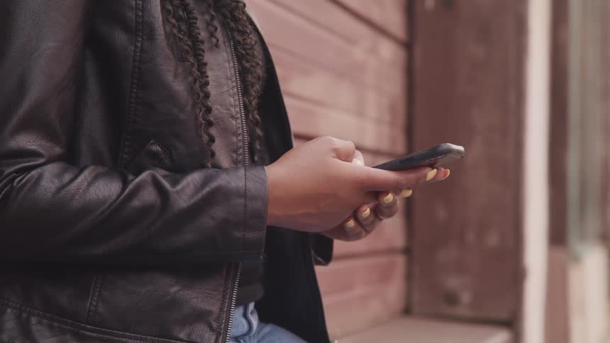 Woman with black leather jacket jeans and braids uses a smartphone in the street