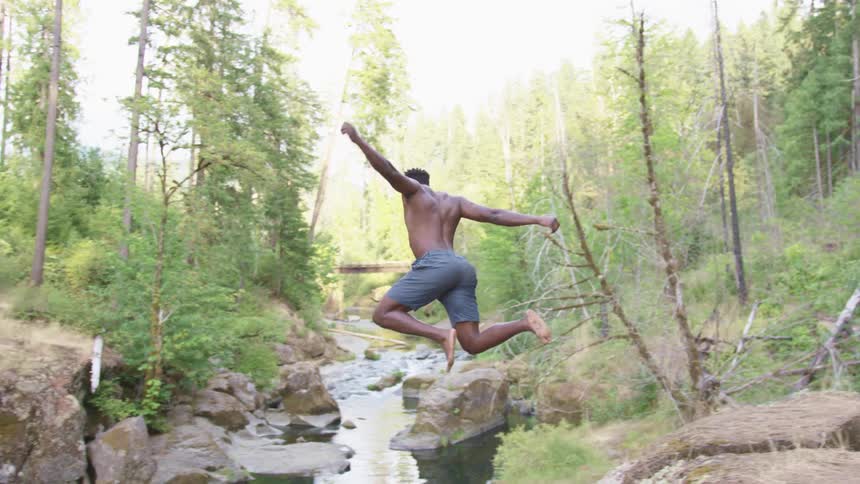 Athletic muscular shirtless man jumping into a river from a hill in the forest