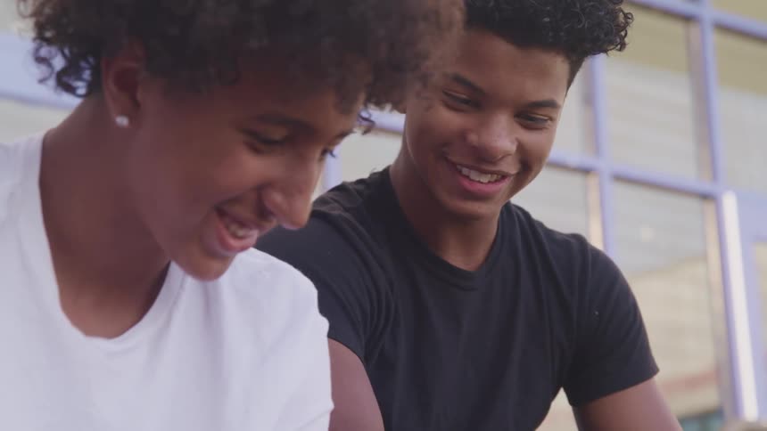 Two friends have fun looking at a phone sitting togheter outside a building