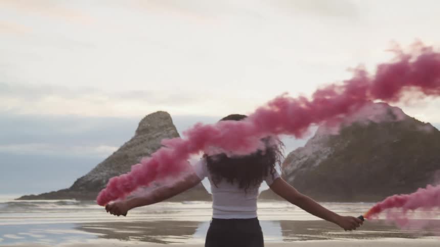 A black female walking on a beach while carrying containers that are spreading red smoke into the air