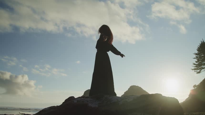Woman stands on rocks near shore stretches her arms out