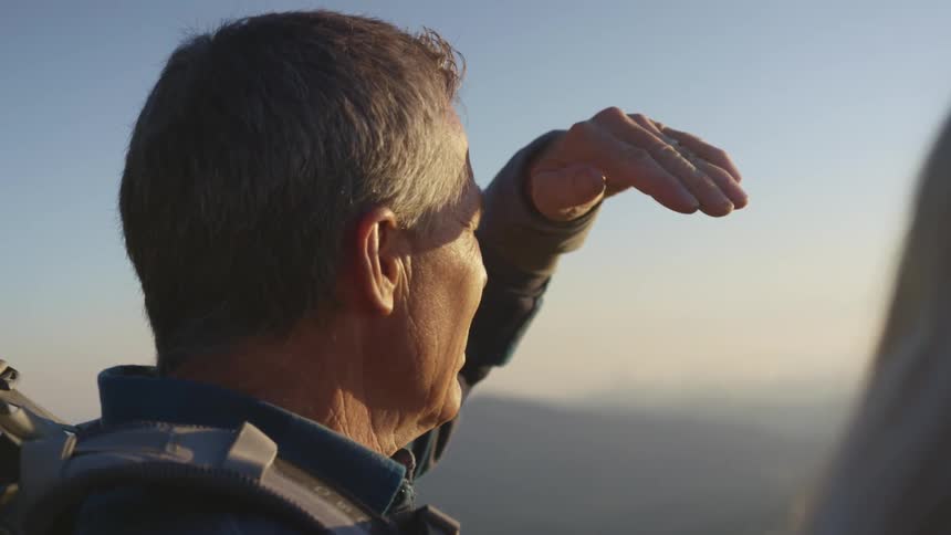A senior citizen hiking on a mountain shielding his eyes with a hand to look at the sunset