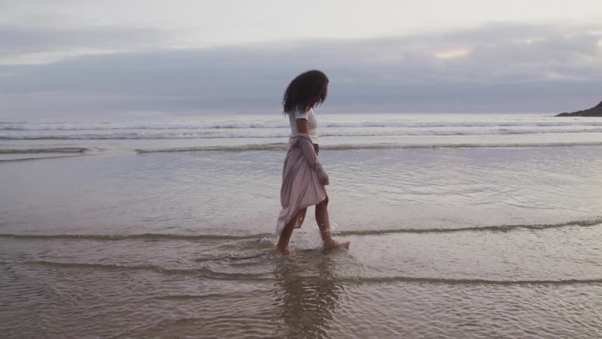 A barefoot woman walking on a wet sandy beach at sunset