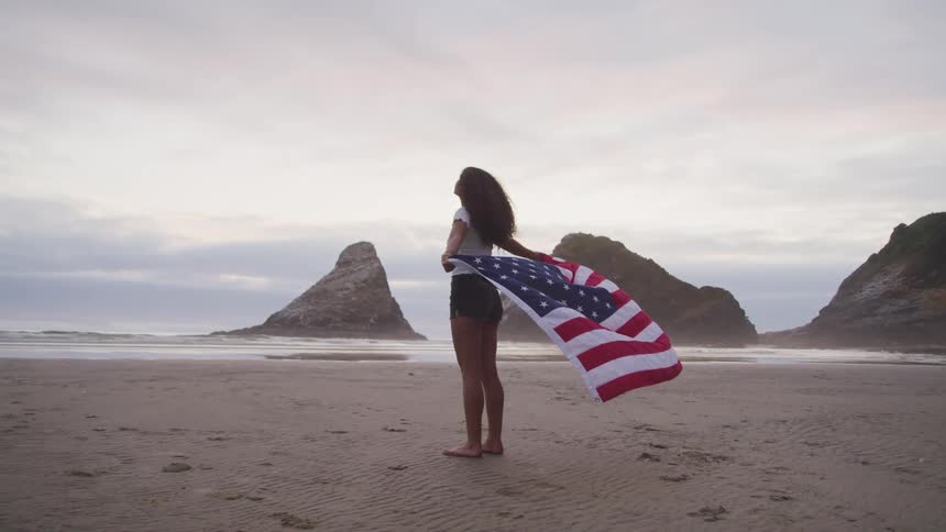 A barefoot woman stands on a beach holding a flag wide behind her
